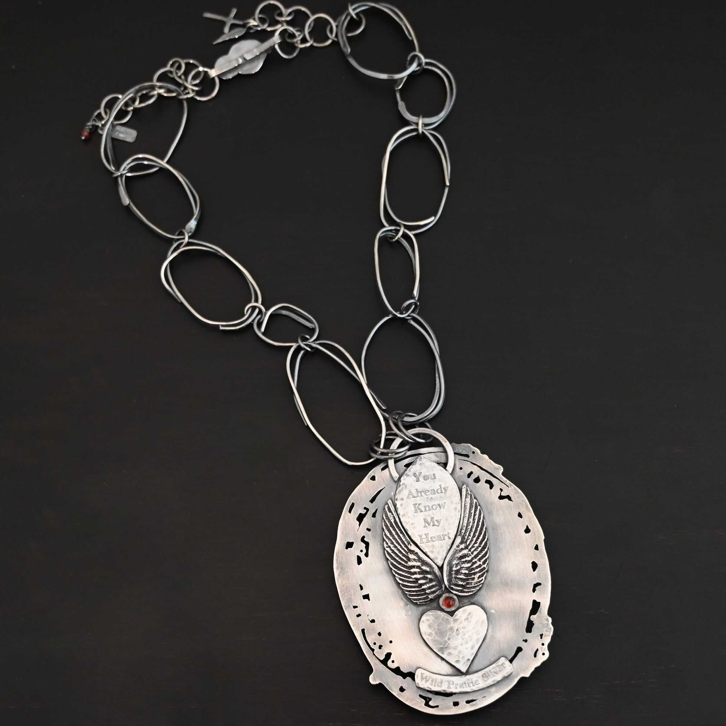 Back of the "Back To Our Roots" Necklace. There is a loop at the top and a marquise shape beneath it with the words, "You already know my heart" stamped into it. There is an angel wing on each side of the marquise shape. There is a small garnet in between the angel wings on the bottom. Beneath this is a hammered heart. Beneath that is a curved piece that says, "Wild Prairie Silver."