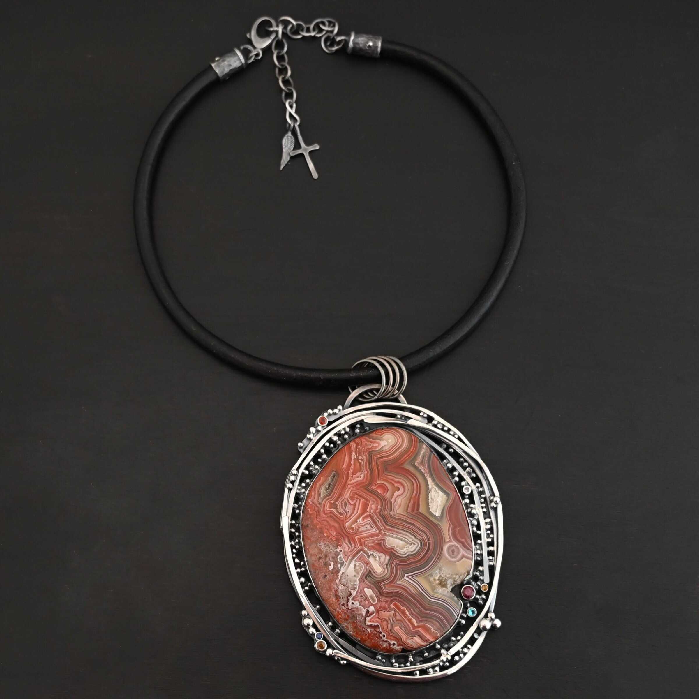 "Back To Our Roots" Necklace. There is a very large oval crazy lace agate in the center with multiple lines of sterling silver wrapped around it. There are small stones scattered around the pendant, along with lots of sterling silver ball detail. Chain is a thick leather cord.