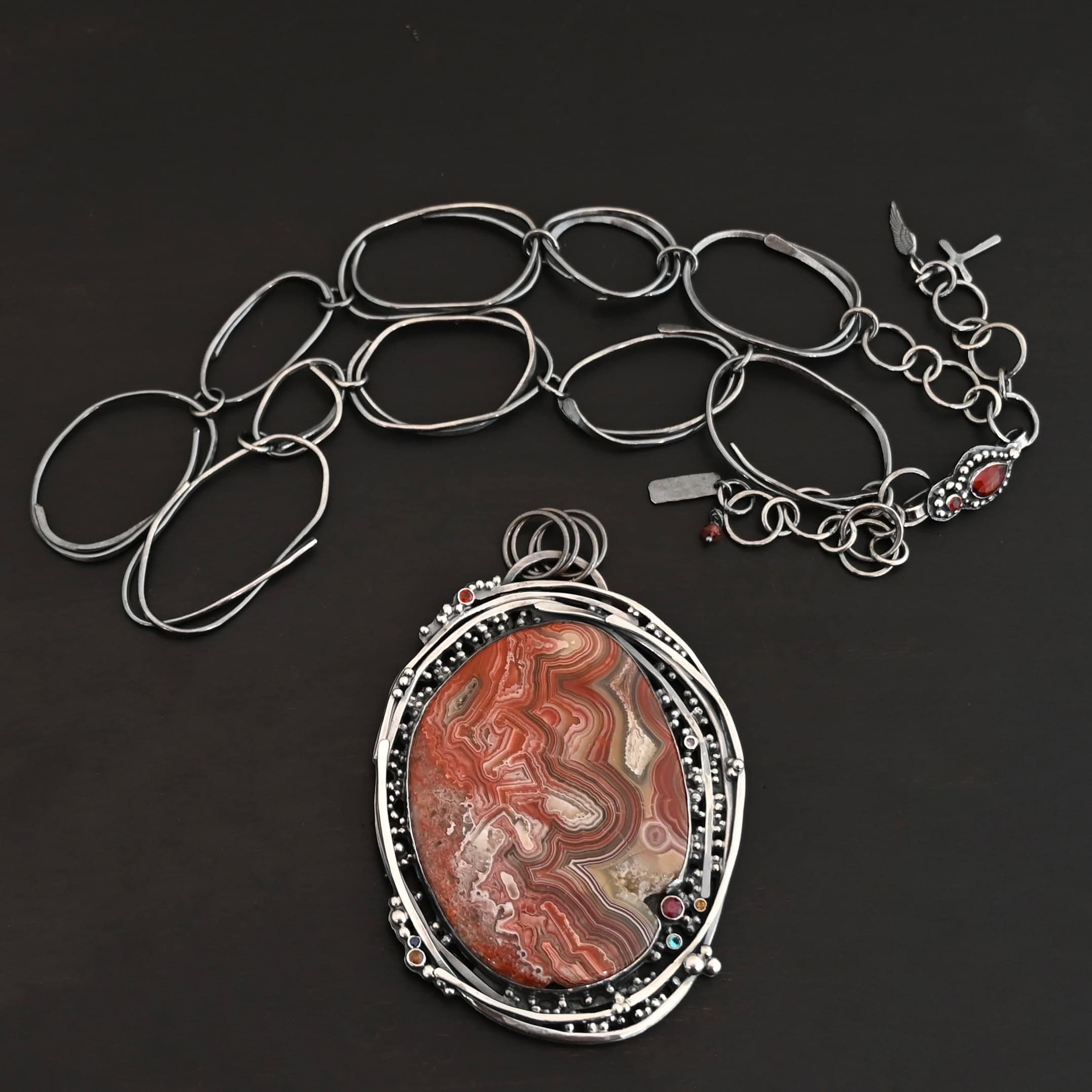 "Back To Our Roots" Necklace. There is a very large oval crazy lace agate in the center with multiple lines of sterling silver wrapped around it. There are small stones scattered around the pendant, along with lots of sterling silver ball detail. Chain is handmade out of large oval links that can detach from the pendant.