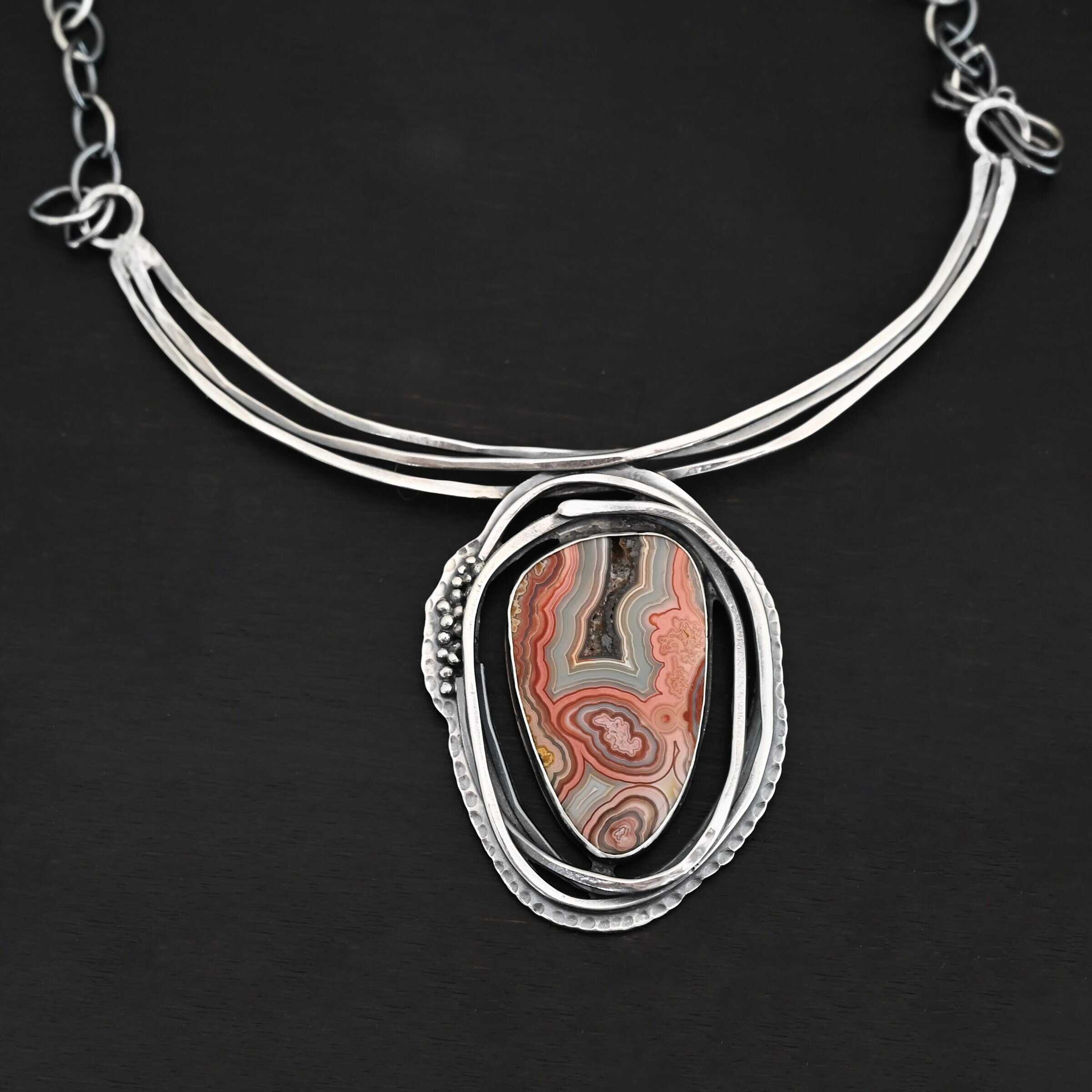 "Try Something New" Necklace. There is a medium sized, oval crazy lace agate in the center. There are multiple lines of silver wrapping around it, but not touching the stone. There is hammered silver and silver ball detail. Pendant is soldered to three large, curved bars of silver that sit around the throat.
