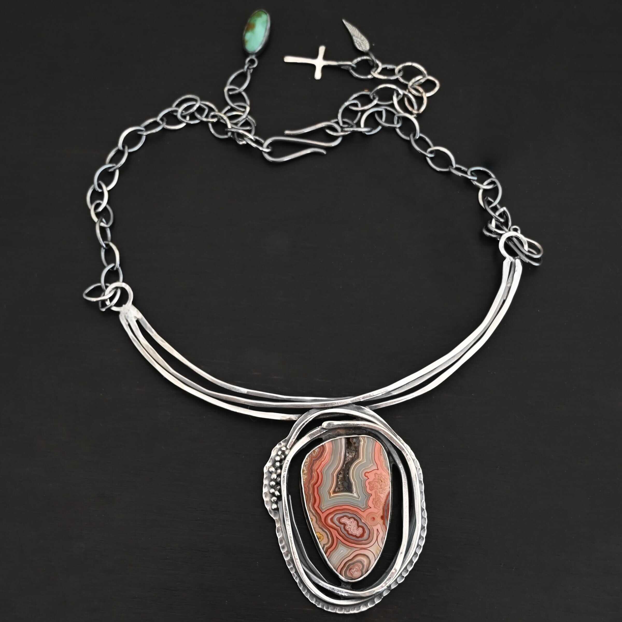 "Try Something New" Necklace. There is a medium sized, oval crazy lace agate in the center. There are multiple lines of silver wrapping around it, but not touching the stone. There is hammered silver and silver ball detail. Pendant is soldered to three large, curved bars of silver that sit around the throat.