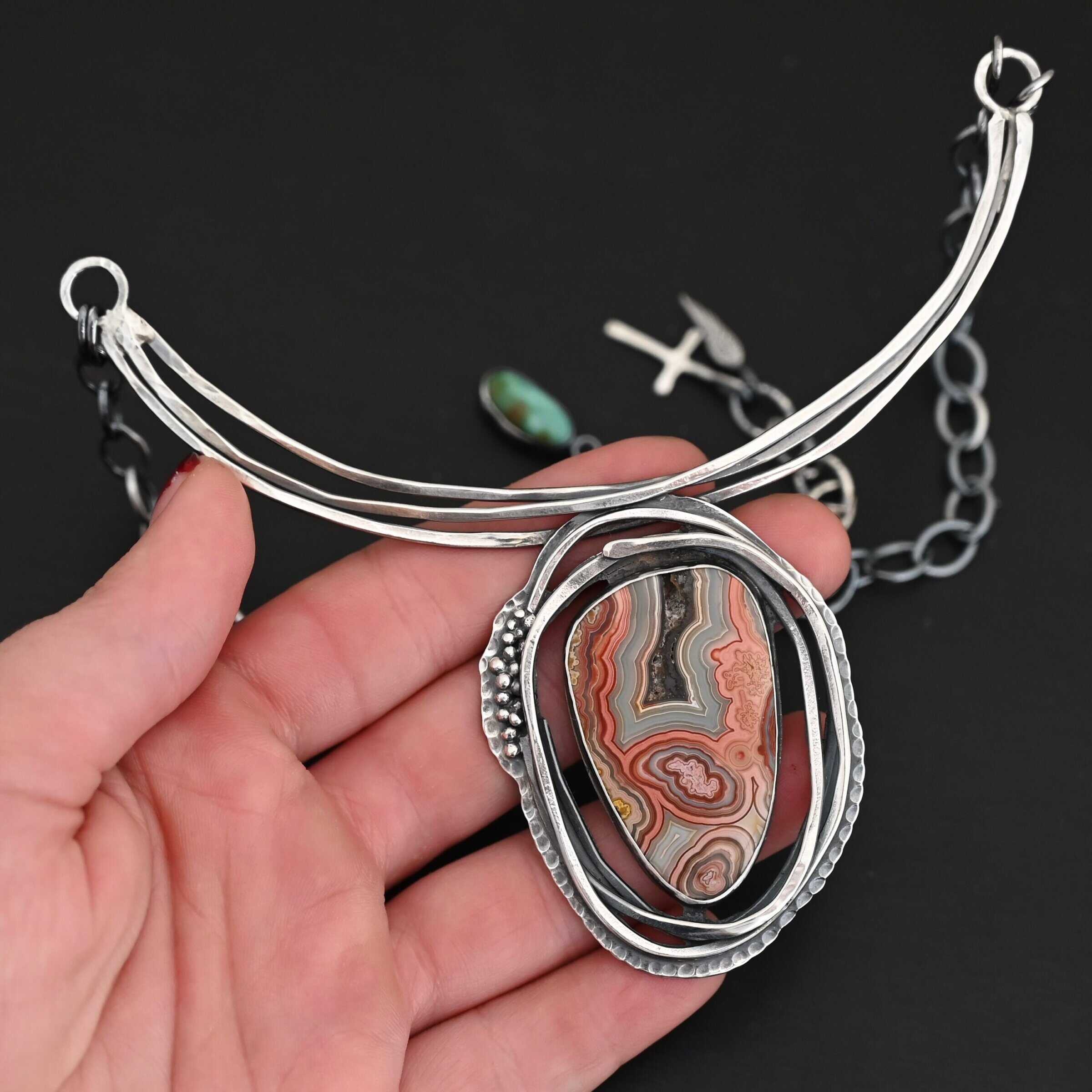 "Try Something New" Necklace. There is a medium sized, oval crazy lace agate in the center. There are multiple lines of silver wrapping around it, but not touching the stone. There is hammered silver and silver ball detail. Pendant is soldered to three large, curved bars of silver that sit around the throat.