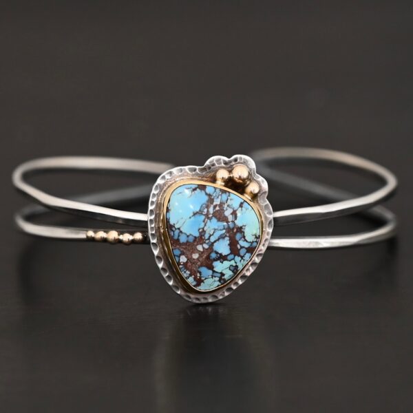"Blue Sky Above" Cuff. There is a medium sized, circular golden hills turquoise in the center. It is set in gold and there are three balls of gold on the top of it. There is hammered silver around the rest of it. The cuff is two separate lines of silver that meet at the back and leave the center empty. There are five balls of gold on the bottom left line.
