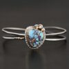 "Blue Sky Above" Cuff. There is a medium sized, circular golden hills turquoise in the center. It is set in gold and there are three balls of gold on the top of it. There is hammered silver around the rest of it. The cuff is two separate lines of silver that meet at the back and leave the center empty. There are five balls of gold on the bottom left line.