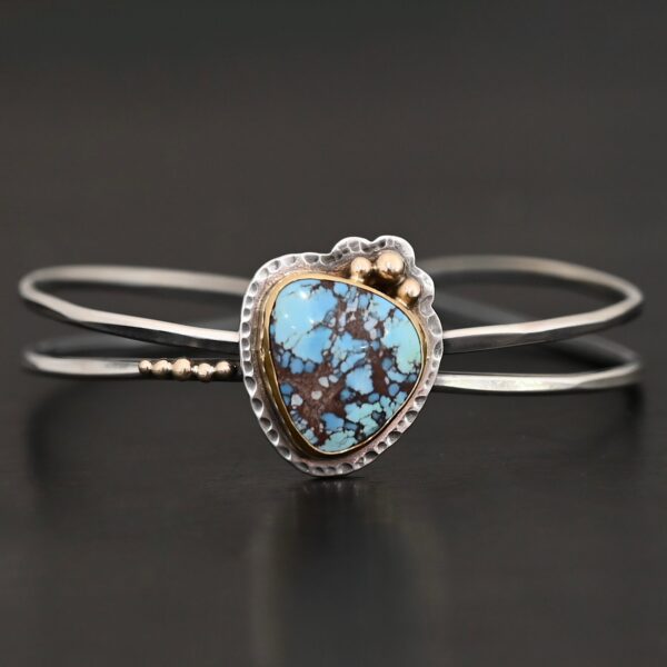 "Blue Sky Above" Cuff. There is a medium sized, circular golden hills turquoise in the center. It is set in gold and there are three balls of gold on the top of it. There is hammered silver around the rest of it. The cuff is two separate lines of silver that meet at the back and leave the center empty. There are five balls of gold on the bottom left line.