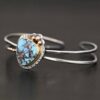 "Blue Sky Above" Cuff. There is a medium sized, circular golden hills turquoise in the center. It is set in gold and there are three balls of gold on the top of it. There is hammered silver around the rest of it. The cuff is two separate lines of silver that meet at the back and leave the center empty. There are five balls of gold on the bottom left line.