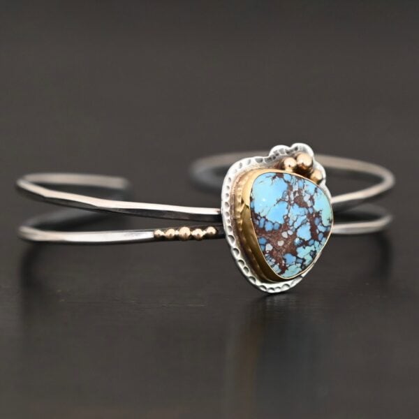 "Blue Sky Above" Cuff. There is a medium sized, circular golden hills turquoise in the center. It is set in gold and there are three balls of gold on the top of it. There is hammered silver around the rest of it. The cuff is two separate lines of silver that meet at the back and leave the center empty. There are five balls of gold on the bottom left line.