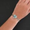 "Blue Sky Above" Cuff. There is a medium sized, circular golden hills turquoise in the center. It is set in gold and there are three balls of gold on the top of it. There is hammered silver around the rest of it. The cuff is two separate lines of silver that meet at the back and leave the center empty. There are five balls of gold on the bottom left line. Model is wearing it up close.