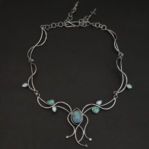 "Siren Song" Necklace. The chain is handmade out of sterling silver. Each link is two lines of silver, one of which is slightly wavy and the other that is very wavy across the top of it. They lead down to an oval vertical blue/green opal. There are four wavy lines of silver that hang around the opal. There are three stones on the end of three of the wavy lines. There are other opals and aquamarines scattered around the chain.