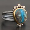 "Kingdom In The Sky" Ring. there is a circular turquoise in the center with three diamonds on the top of it and one on the bottom. There are four balls of gold on top of it, one on each end and two in between, and two balls of gold on the bottom, one on each side of the diamond. Ring band is two lines of silver, one soldered to the top and one to the bottom. They meet at the back of the ring and leave the center empty.