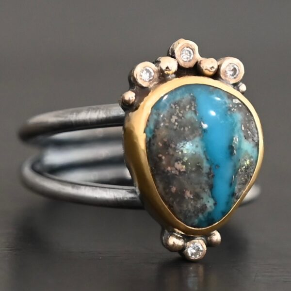 "Kingdom In The Sky" Ring. there is a circular turquoise in the center with three diamonds on the top of it and one on the bottom. There are four balls of gold on top of it, one on each end and two in between, and two balls of gold on the bottom, one on each side of the diamond. Ring band is two lines of silver, one soldered to the top and one to the bottom. They meet at the back of the ring and leave the center empty.