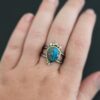 "Kingdom In The Sky" Ring. there is a circular turquoise in the center with three diamonds on the top of it and one on the bottom. There are four balls of gold on top of it, one on each end and two in between, and two balls of gold on the bottom, one on each side of the diamond. Ring band is two lines of silver, one soldered to the top and one to the bottom. They meet at the back of the ring and leave the center empty. Ring is worn up close by a model.