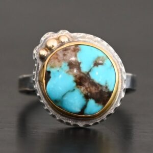 "Kingdom In The Sky" Ring. There is a circular turquoise in the center with three balls of gold on the top left side of it. There is hammered silver around the rest of it. Ring band is smooth oxidized silver.
