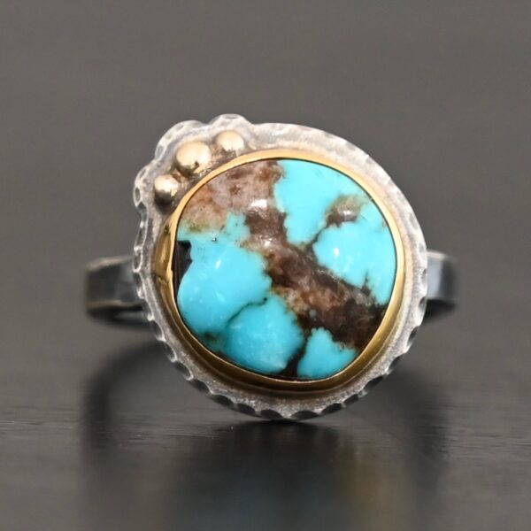 "Kingdom In The Sky" Ring. There is a circular turquoise in the center with three balls of gold on the top left side of it. There is hammered silver around the rest of it. Ring band is smooth oxidized silver.