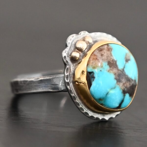 "Kingdom In The Sky" Ring. There is a circular turquoise in the center with three balls of gold on the top left side of it. There is hammered silver around the rest of it. Ring band is smooth oxidized silver.