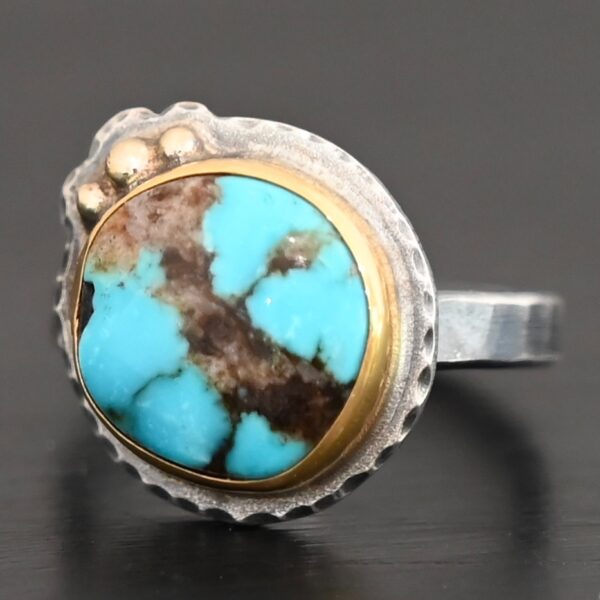 "Kingdom In The Sky" Ring. There is a circular turquoise in the center with three balls of gold on the top left side of it. There is hammered silver around the rest of it. Ring band is smooth oxidized silver.