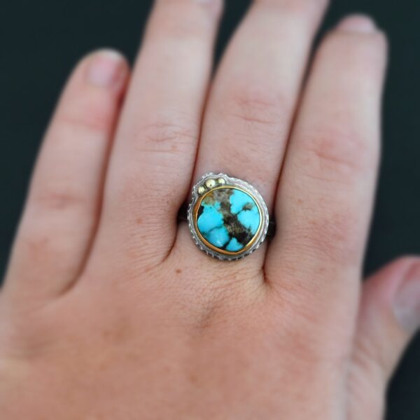 "Kingdom In The Sky" Ring. There is a circular turquoise in the center with three balls of gold on the top left side of it. There is hammered silver around the rest of it. Ring band is smooth oxidized silver. Ring is shown up close by a model.