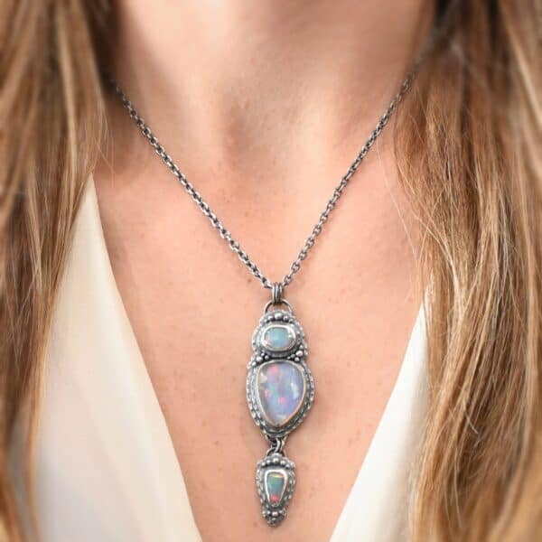 "Blue Moon Magic" Necklace. There are three opals in a line down. The top one is a small horizontal oval. The next one is a larger, vertical opal. The bottom one is a small triangle. Each one has silver ball detail surrounding it, as well as hammered silver. Necklace is shown up close by a model wearing it.