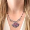 "In The Dawns" Necklace. There are six rubies and one large, wide, pear shaped red/pink opal pointing up. The opal is at the bottom, with five oval vertical rubies circling around the bottom of it. In between each ruby is a silver leaf and on the end of each ruby is a ball of silver. The clasp of the necklace has three silver leaves and a ball of silver above an oval ruby. Chain is handmade out of wavy links of sterling silver.