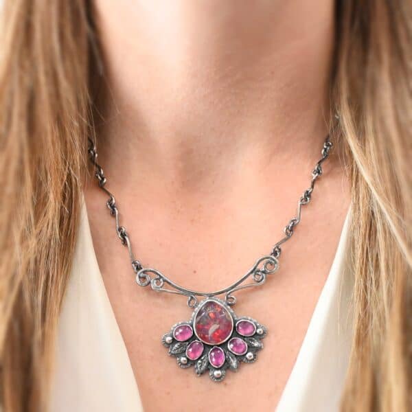 "In The Dawns" Necklace. There are six rubies and one large, wide, pear shaped red/pink opal pointing up. The opal is at the bottom, with five oval vertical rubies circling around the bottom of it. In between each ruby is a silver leaf and on the end of each ruby is a ball of silver. The clasp of the necklace has three silver leaves and a ball of silver above an oval ruby. Chain is handmade out of wavy links of sterling silver.