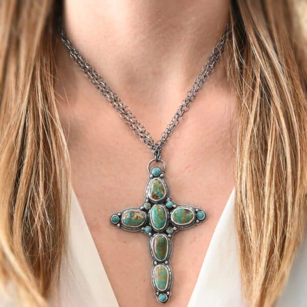 "Firm In Faith" Necklace. Necklace is a large cross shape with six oval turquoise creating the cross shape and eight small circular turquoise around the rest of the pendant. Chain is multiple strands that meet at the back. Necklace is shown up close by a model.
