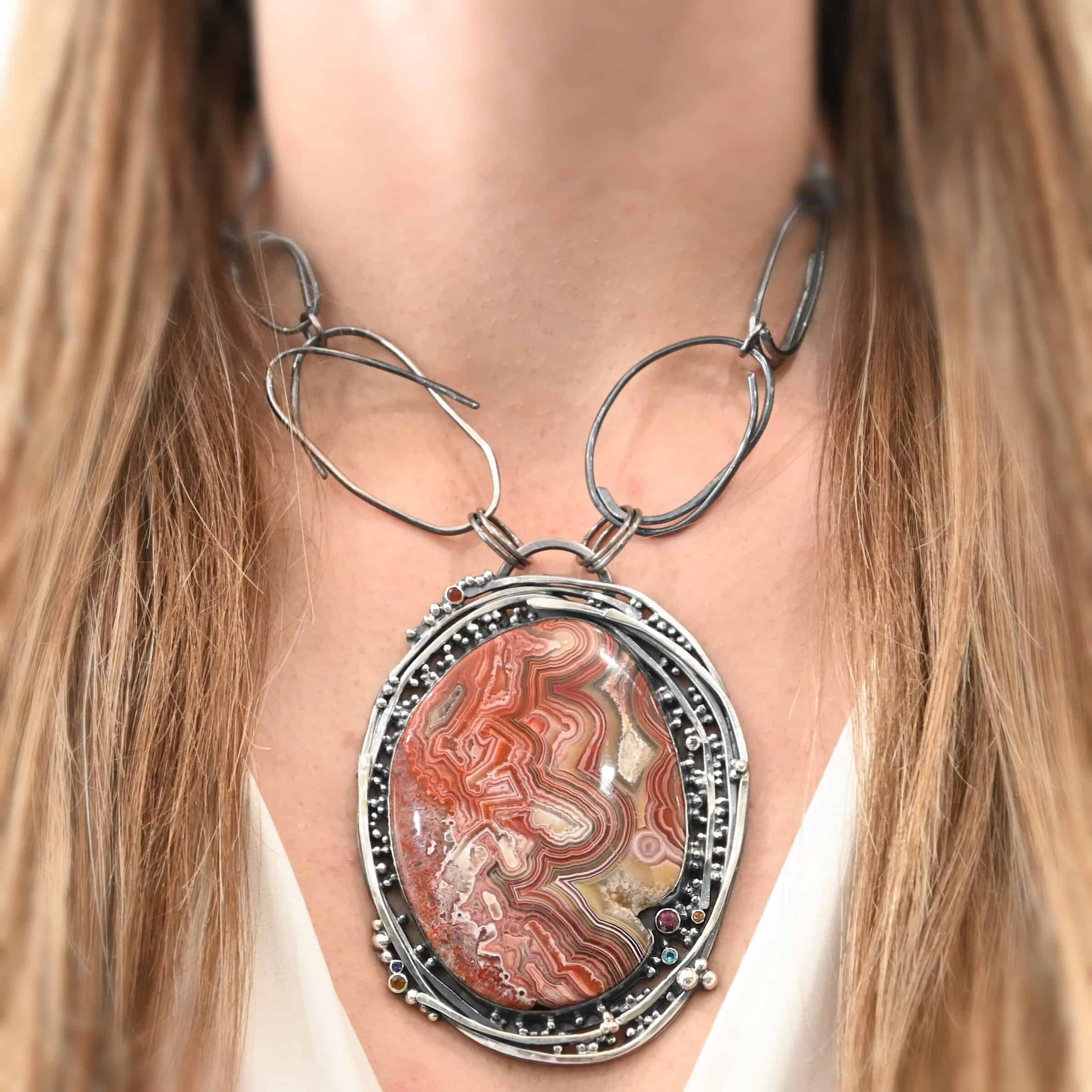 "Back To Our Roots" Necklace. There is a very large oval crazy lace agate in the center with multiple lines of sterling silver wrapped around it. There are small stones scattered around the pendant, along with lots of sterling silver ball detail. Chain is handmade out of large oval links that can detach from the pendant. Necklace is shown up close by a model.