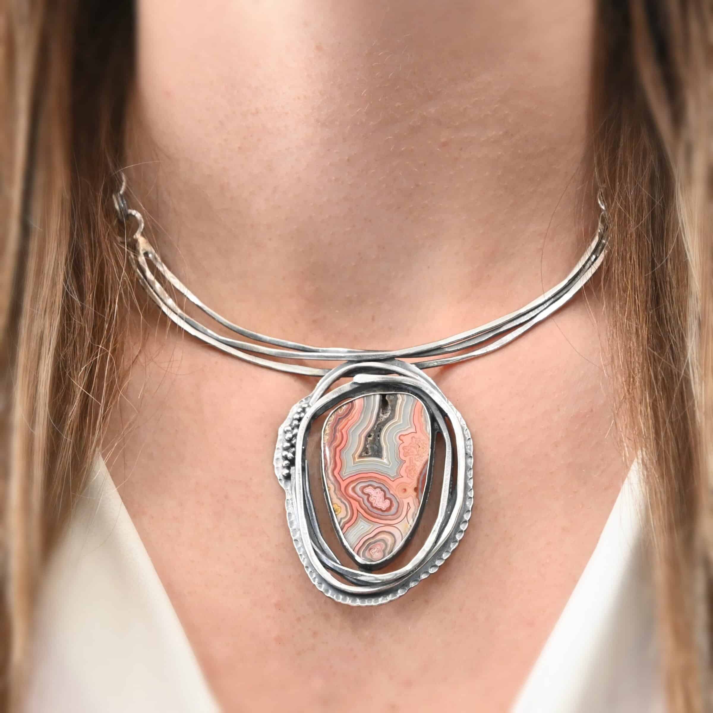 "Try Something New" Necklace. There is a medium sized, oval crazy lace agate in the center. There are multiple lines of silver wrapping around it, but not touching the stone. There is hammered silver and silver ball detail. Pendant is soldered to three large, curved bars of silver that sit around the throat.