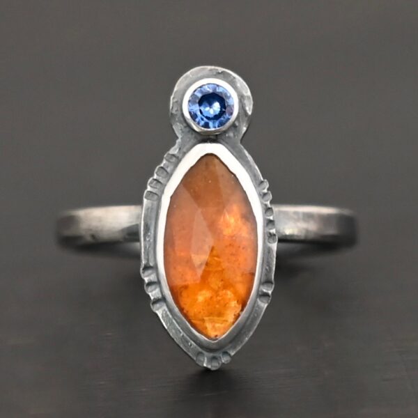 Photo of "The Sun Will Rise" Ring. Made out of sterling silver, natural orange kyanite, and tourmaline. There is a thin silver band, with a marquise shaped orange kyanite in the center, and a small, circular, cornflower blue tourmaline on the top tip of the marquise. Ring is sitting against a dark background.