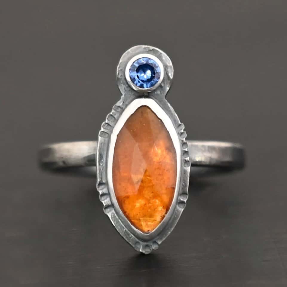 The Sun Will Rise Ring Photo of "The Sun Will Rise" Ring. Made out of sterling silver, natural orange kyanite, and tourmaline. There is a thin silver band, with a marquise shaped orange kyanite in the center, and a small, circular, cornflower blue tourmaline on the top tip of the marquise. Ring is sitting against a dark background.