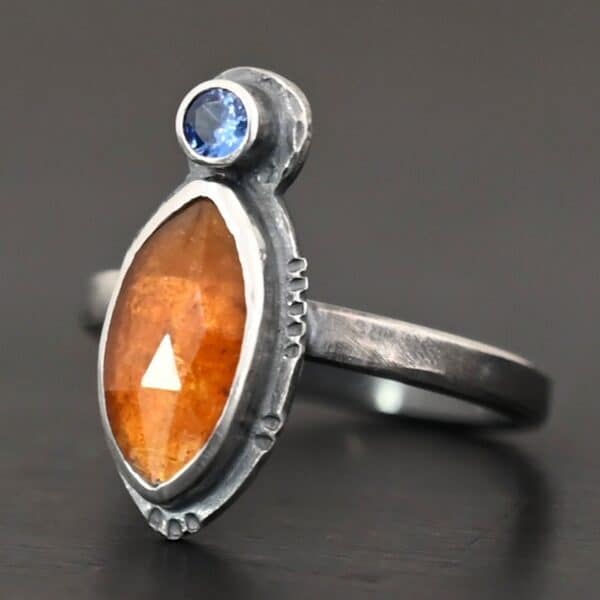 Photo of "The Sun Will Rise" Ring. Made out of sterling silver, natural orange kyanite, and tourmaline. There is a thin silver band, with a marquise shaped orange kyanite in the center, and a small, circular, cornflower blue tourmaline on the top tip of the marquise. Ring is sitting against a dark background.