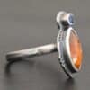 Photo of "The Sun Will Rise" Ring. Made out of sterling silver, natural orange kyanite, and tourmaline. There is a thin silver band, with a marquise shaped orange kyanite in the center, and a small, circular, cornflower blue tourmaline on the top tip of the marquise. Ring is sitting against a dark background.