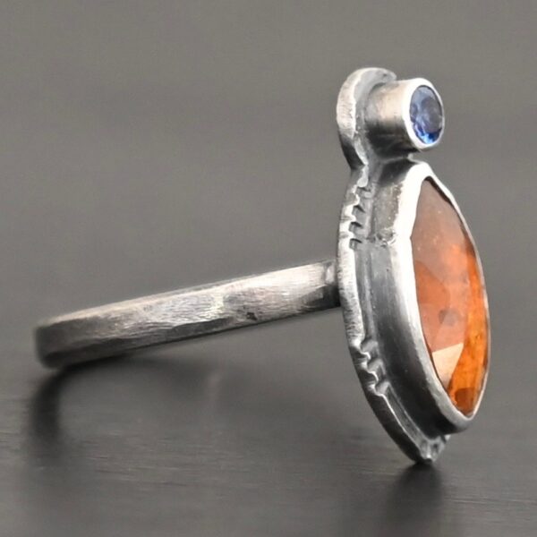 Photo of "The Sun Will Rise" Ring. Made out of sterling silver, natural orange kyanite, and tourmaline. There is a thin silver band, with a marquise shaped orange kyanite in the center, and a small, circular, cornflower blue tourmaline on the top tip of the marquise. Ring is sitting against a dark background.