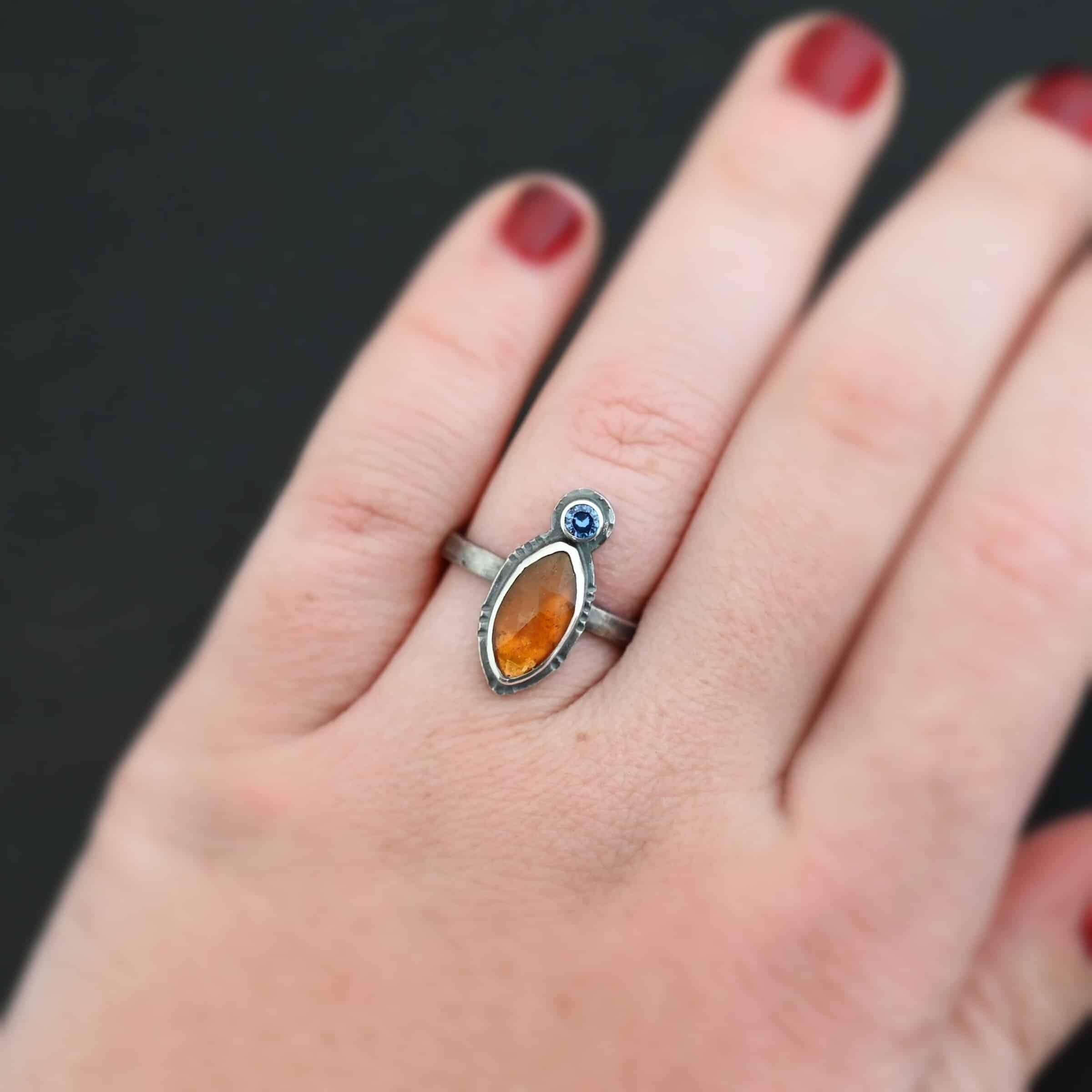 The Sun Will Rise Ring Photo of "The Sun Will Rise" Ring. Made out of sterling silver, natural orange kyanite, and tourmaline. There is a thin silver band, with a marquise shaped orange kyanite in the center, and a small, circular, cornflower blue tourmaline on the top tip of the marquise. Ring is worn by a model to demonstrate size.