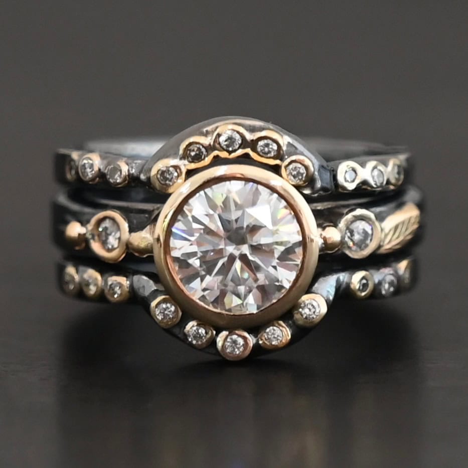 "In Another Life" Ring. There is a round, two carat moissanite in the center of the ring. There are two smaller moissanites, one on each side of it. There are two more bands, one on the top and bottom of the ring. Each has multiple moissanites that go around it. There is gold ball detail and gold leaves on the center band as well.