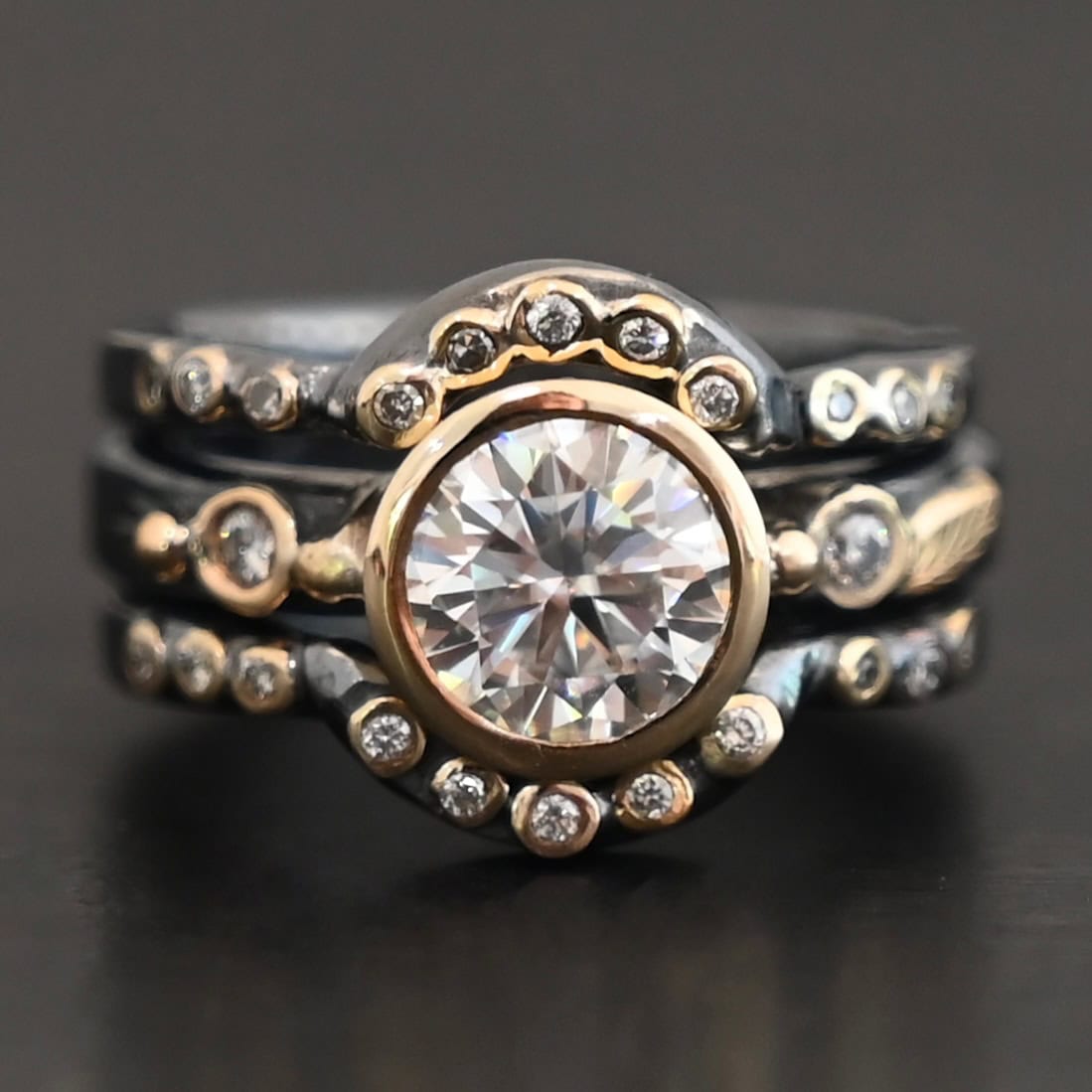"In Another Life" Ring. There is a round, two carat moissanite in the center of the ring. There are two smaller moissanites, one on each side of it. There are two more bands, one on the top and bottom of the ring. Each has multiple moissanites that go around it. There is gold ball detail and gold leaves on the center band as well.