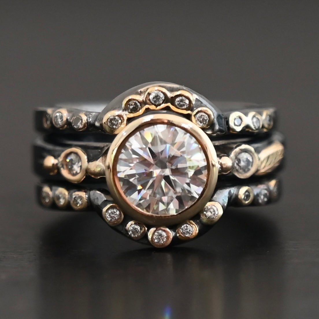 "In Another Life" Ring. There is a round, two carat moissanite in the center of the ring. There are two smaller moissanites, one on each side of it. There are two more bands, one on the top and bottom of the ring. Each has multiple moissanites that go around it. There is gold ball detail and gold leaves on the center band as well.
