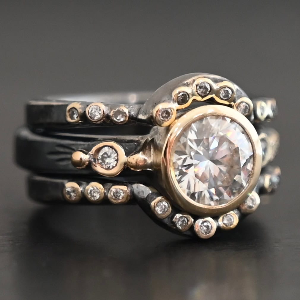 "In Another Life" Ring. There is a round, two carat moissanite in the center of the ring. There are two smaller moissanites, one on each side of it. There are two more bands, one on the top and bottom of the ring. Each has multiple moissanites that go around it. There is gold ball detail and gold leaves on the center band as well.