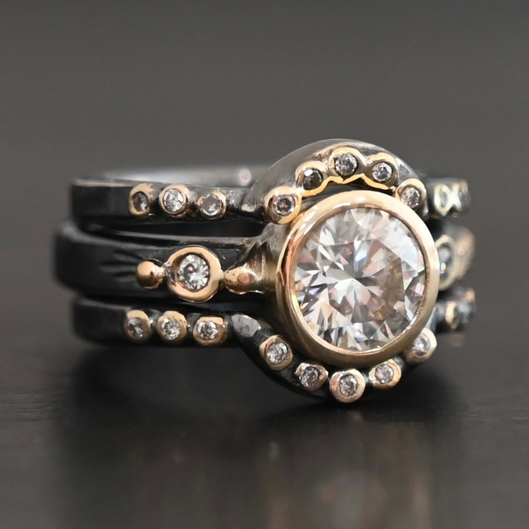 "In Another Life" Ring. There is a round, two carat moissanite in the center of the ring. There are two smaller moissanites, one on each side of it. There are two more bands, one on the top and bottom of the ring. Each has multiple moissanites that go around it. There is gold ball detail and gold leaves on the center band as well.