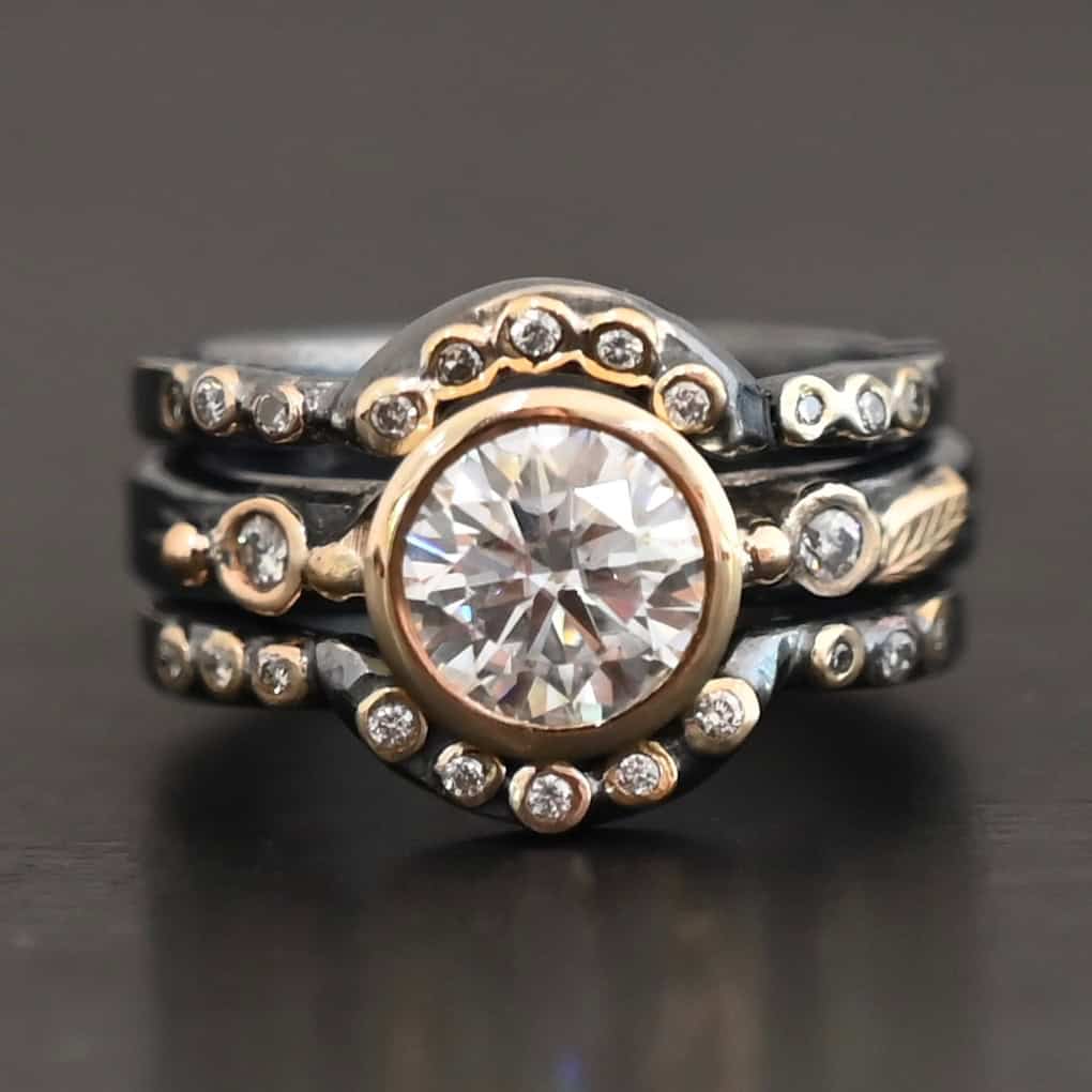 "In Another Life" Ring. There is a round, two carat moissanite in the center of the ring. There are two smaller moissanites, one on each side of it. There are two more bands, one on the top and bottom of the ring. Each has multiple moissanites that go around it. There is gold ball detail and gold leaves on the center band as well.