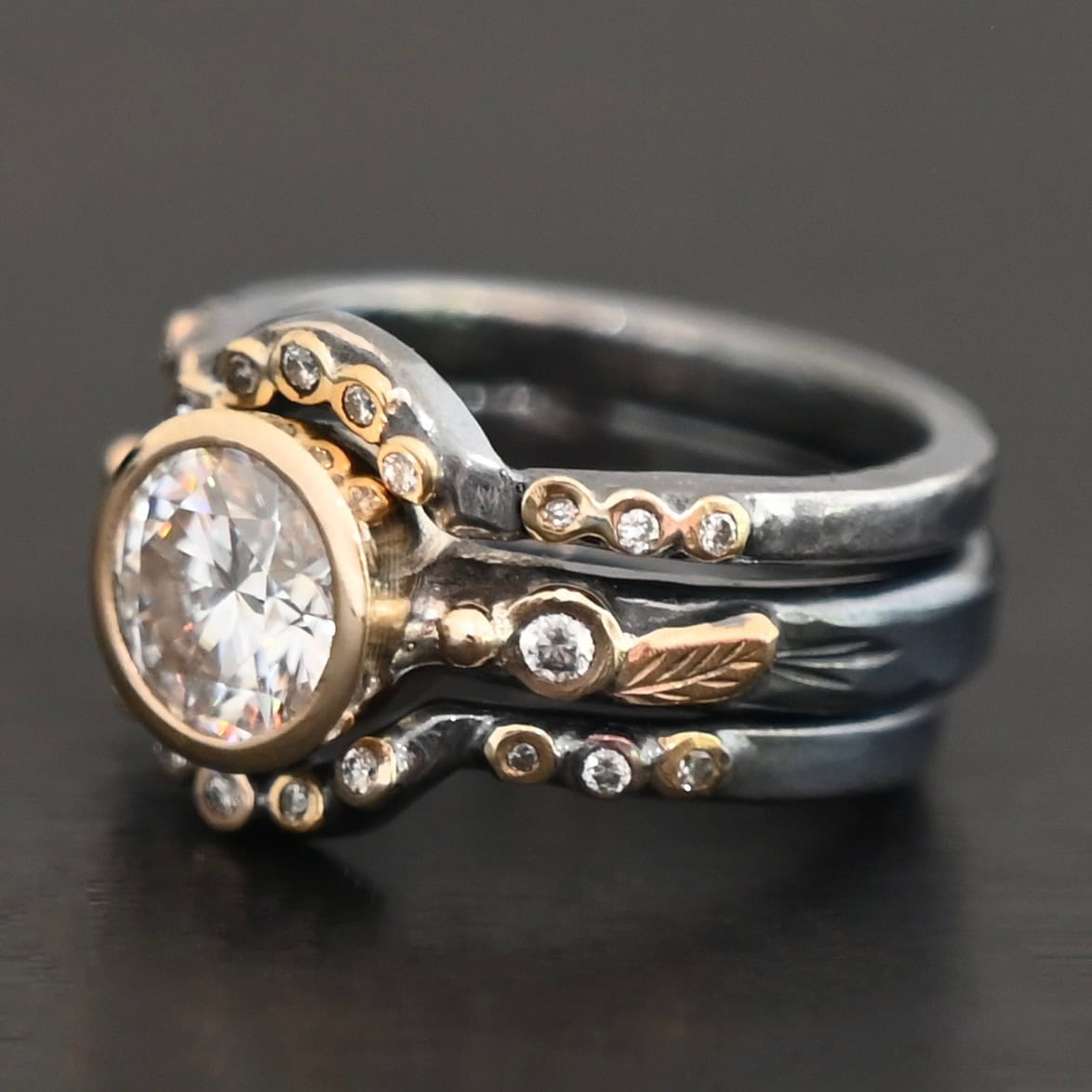 "In Another Life" Ring. There is a round, two carat moissanite in the center of the ring. There are two smaller moissanites, one on each side of it. There are two more bands, one on the top and bottom of the ring. Each has multiple moissanites that go around it. There is gold ball detail and gold leaves on the center band as well.
