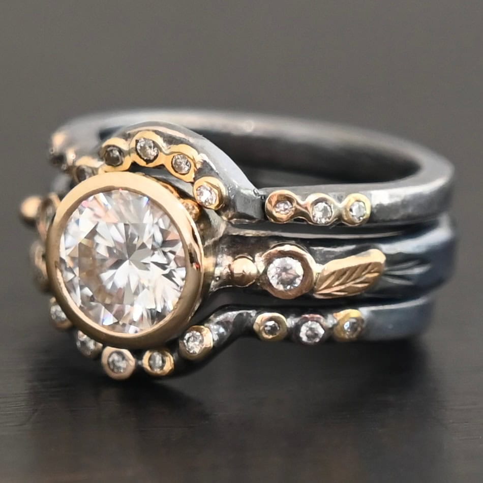 "In Another Life" Ring. There is a round, two carat moissanite in the center of the ring. There are two smaller moissanites, one on each side of it. There are two more bands, one on the top and bottom of the ring. Each has multiple moissanites that go around it. There is gold ball detail and gold leaves on the center band as well.