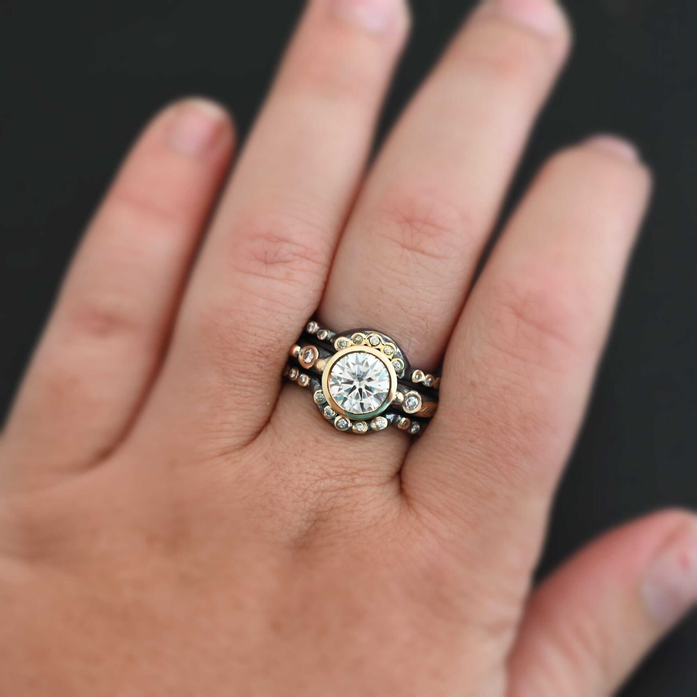 "In Another Life" Ring. There is a round, two carat moissanite in the center of the ring. There are two smaller moissanites, one on each side of it. There are two more bands, one on the top and bottom of the ring. Each has multiple moissanites that go around it. There is gold ball detail and gold leaves on the center band as well. Ring is shown up close by a model on their middle finger to demonstrate size.