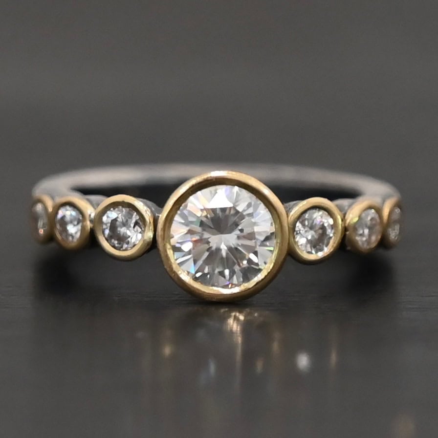 "Diana" Ring. There is two carat moissanite in the center of the ring, with six more smaller moissanites, three on each side of the center one. Each stone is set in gold. The ring band is silver.