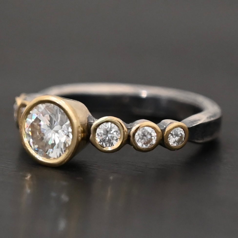 "Diana" Ring. There is two carat moissanite in the center of the ring, with six more smaller moissanites, three on each side of the center one. Each stone is set in gold. The ring band is silver.