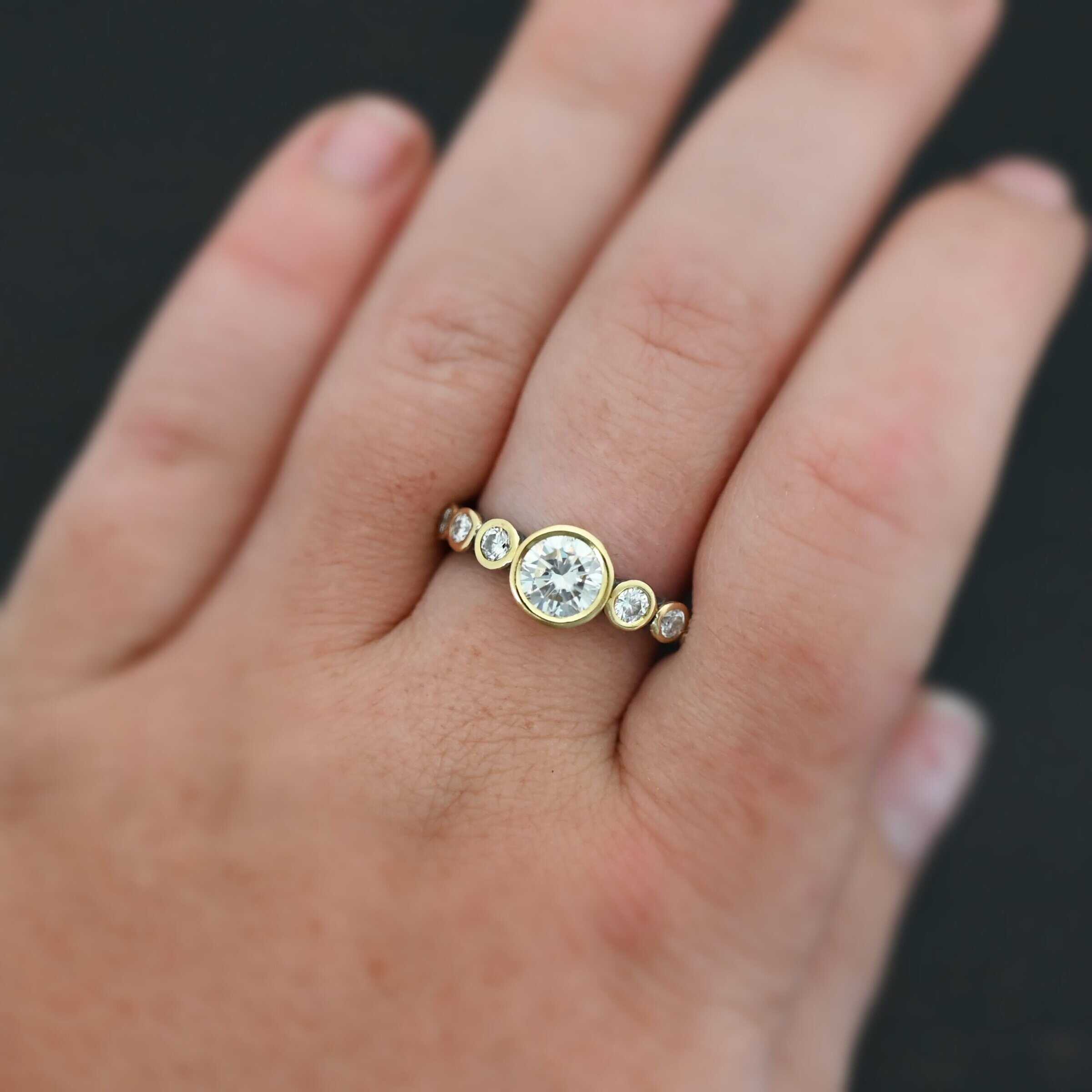 "Diana" Ring. There is two carat moissanite in the center of the ring, with six more smaller moissanites, three on each side of the center one. Each stone is set in gold. The ring band is silver. Ring is shown up close by a model on their middle finger to demonstrate size.