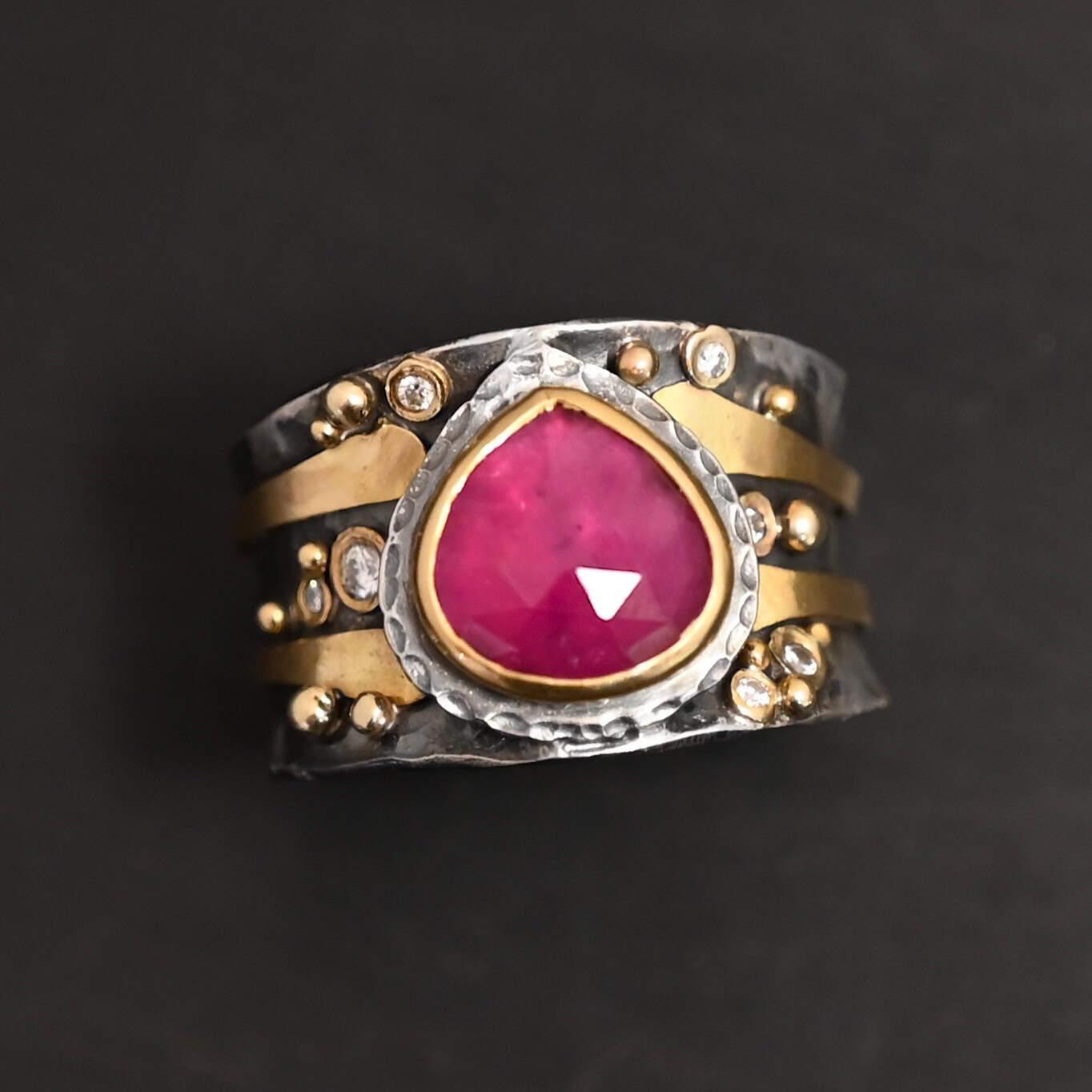 "Cherry Tree" Ring. Ring band is wide and sterling silver. There is a wide, pear shaped ruby in the center with hammered silver around it. There are two thicker bands of gold that wrap around the band. There are small diamonds and balls of gold scattered around the rest of the ring band.
