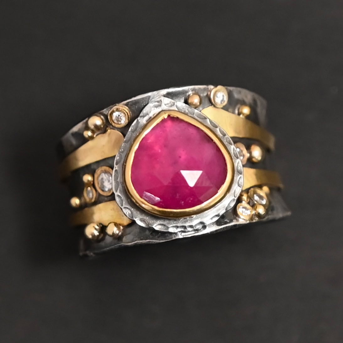 "Cherry Tree" Ring. Ring band is wide and sterling silver. There is a wide, pear shaped ruby in the center with hammered silver around it. There are two thicker bands of gold that wrap around the band. There are small diamonds and balls of gold scattered around the rest of the ring band.