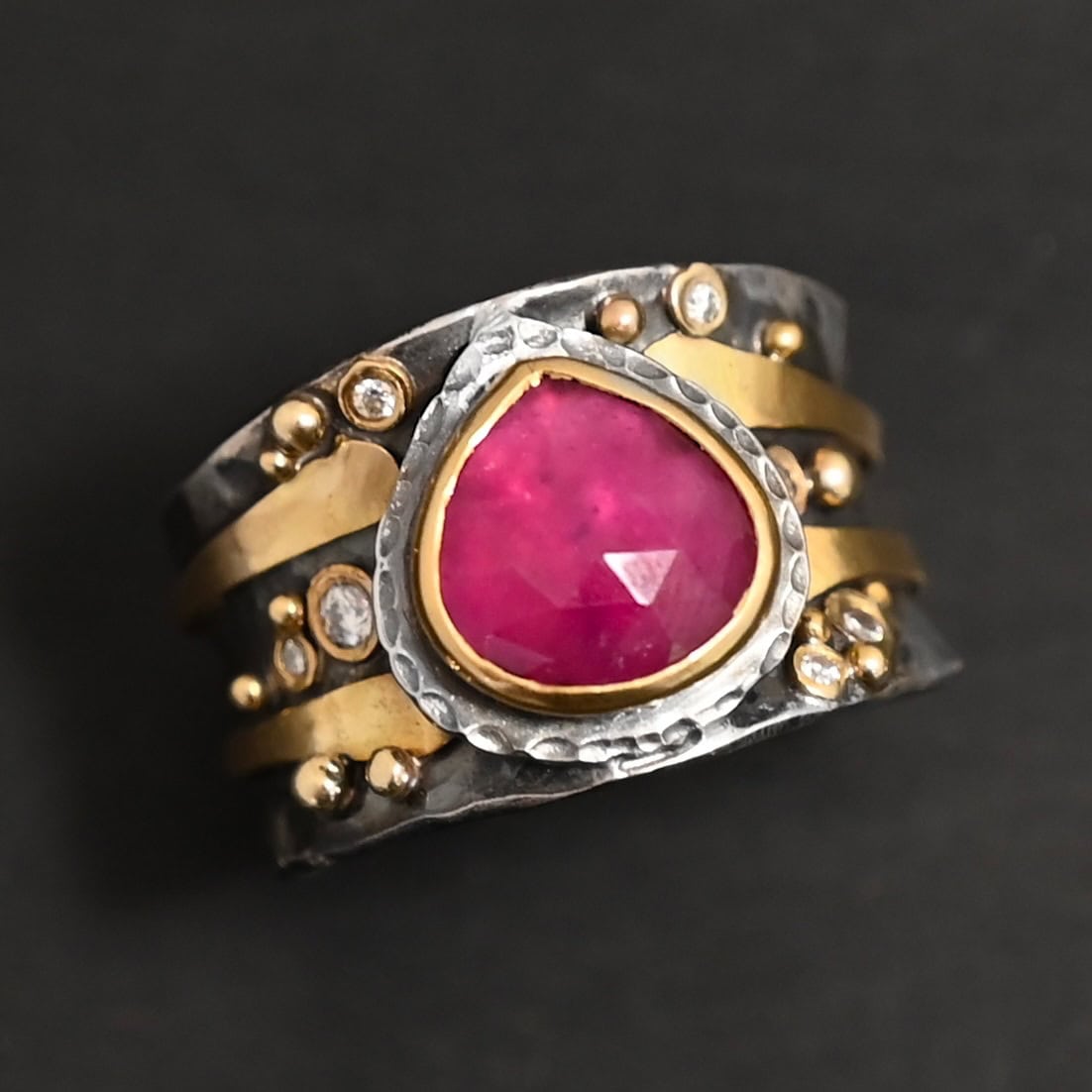 "Cherry Tree" Ring. Ring band is wide and sterling silver. There is a wide, pear shaped ruby in the center with hammered silver around it. There are two thicker bands of gold that wrap around the band. There are small diamonds and balls of gold scattered around the rest of the ring band.
