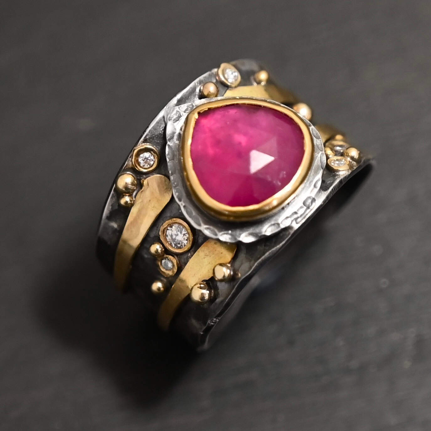 "Cherry Tree" Ring. Ring band is wide and sterling silver. There is a wide, pear shaped ruby in the center with hammered silver around it. There are two thicker bands of gold that wrap around the band. There are small diamonds and balls of gold scattered around the rest of the ring band.