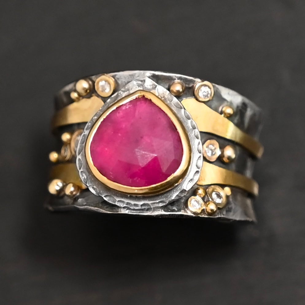 "Cherry Tree" Ring. Ring band is wide and sterling silver. There is a wide, pear shaped ruby in the center with hammered silver around it. There are two thicker bands of gold that wrap around the band. There are small diamonds and balls of gold scattered around the rest of the ring band.